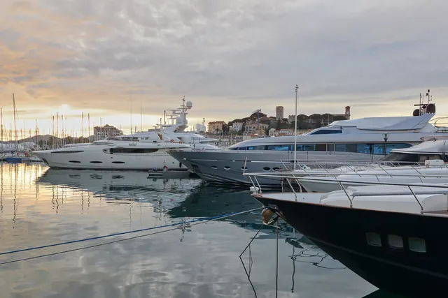 Yachts amarrés au Vieux-Port de Cannes au crépuscule, avec la proue sombre d'un bateau au premier plan et, au fond, la colline du Suquet, sa tour et son clocher, sous un ciel doré à gauche.