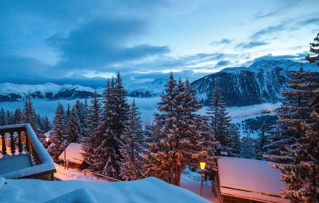 Chalets et sapins enneigés de Courchevel à l'heure bleue, avec un lampadaire allumé, vus depuis un balcon ; mer de nuages et sommets enneigés au loin.