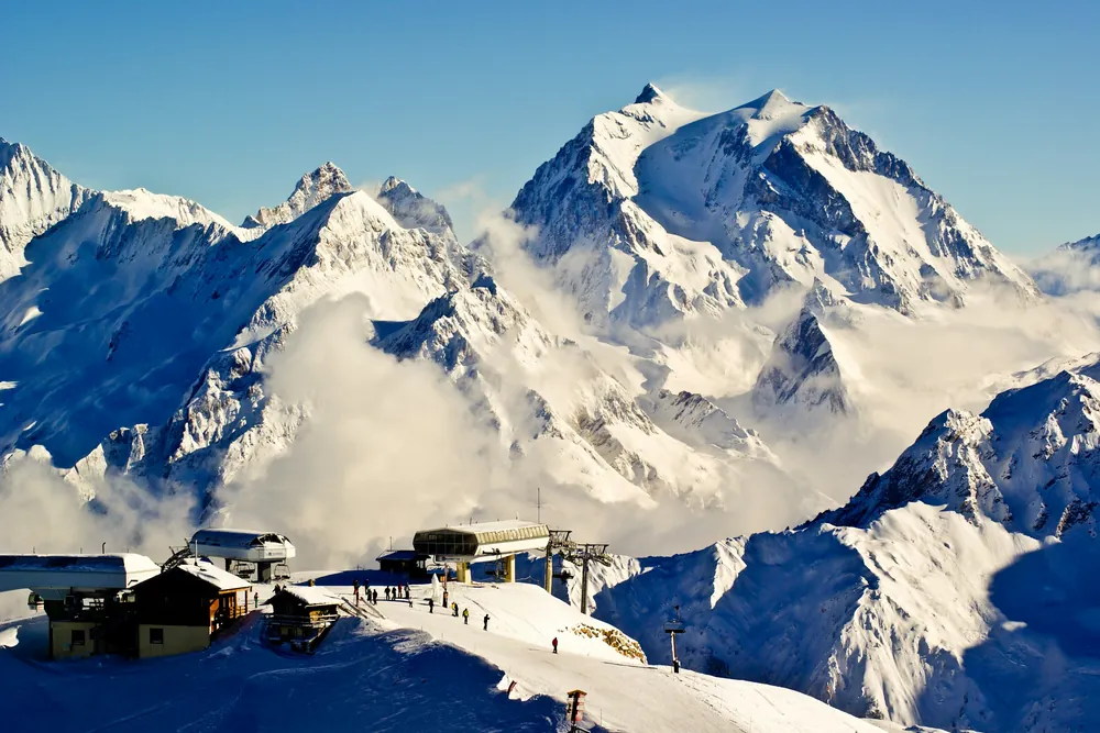Gares de télésiège et chalets au sommet d'un domaine skiable, face à des sommets enneigés à demi noyés dans les nuages.