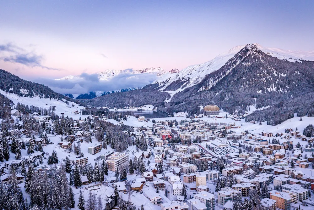 Davos sous la neige au crépuscule, vue du ciel : la station aux toits blancs, le lac gelé au fond de la vallée et les sommets enneigés sous un ciel rose et mauve.