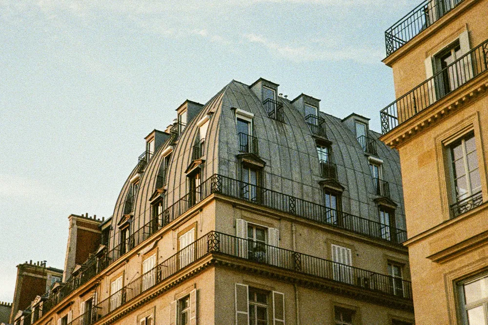 Immeuble haussmannien à l'angle d'une rue de Paris, avec son toit de zinc bombé, ses lucarnes et ses balcons en fer forgé dans la lumière du soir.