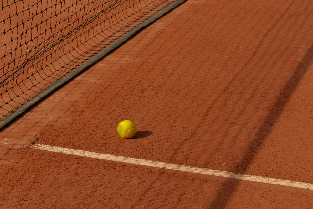 Balle de tennis posée sur la terre battue, près du filet et d'une ligne blanche, dans une lumière rasante.