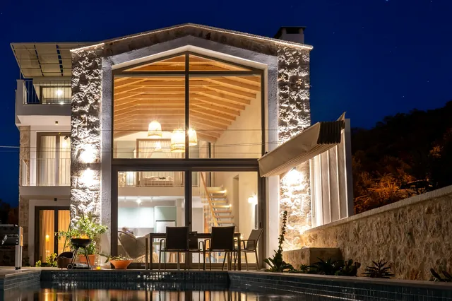 Villa en pierre éclairée de nuit, grande baie vitrée sur un intérieur boisé, terrasse et piscine au premier plan sous un ciel étoilé.